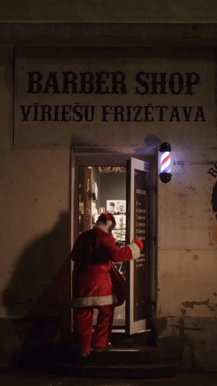 Ieeja bārberšopā vakarā ar izkārtni „Barber shop / Vīriešu frizētava” un bārddziņa stabu
